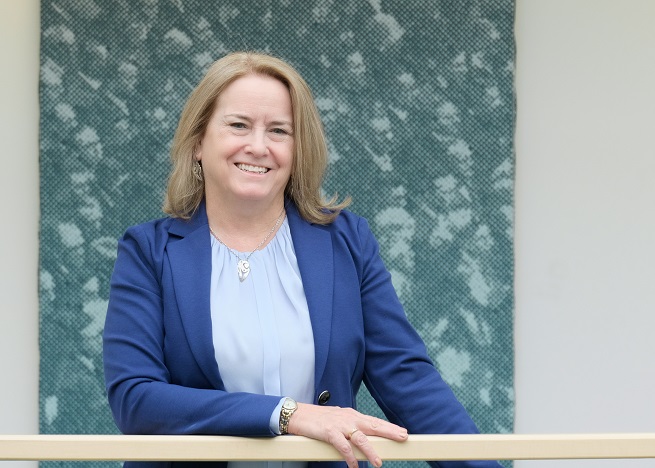 A photo of Dr. Mary Ingraham, Dean of Arts. She is wearing a blue suit and she's standing in front of an abstract artwork on the third level of the Atrium Building.